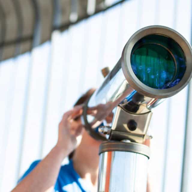 Person looking through a telescope with a blue lens, set against a backdrop of a sunny sky and the metal structure of CommissionVue's sales commission management headquarters.