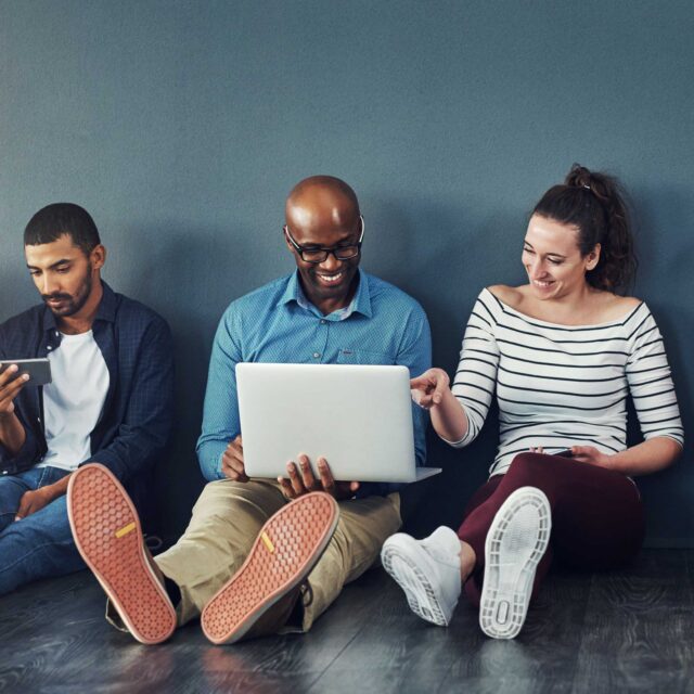 Three people sitting against a wall, using electronic devices. The man in the middle is laughing and sharing his SolutionVue laptop screen with a woman next to him; another man is focused on his phone.