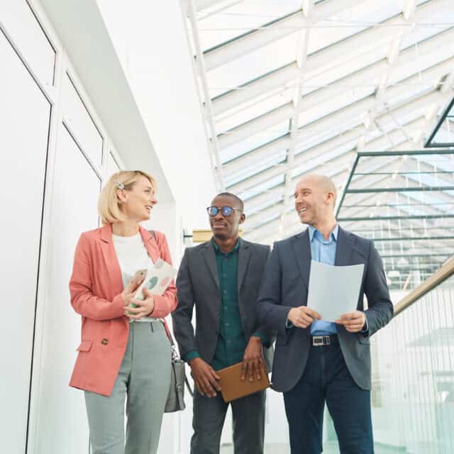 Three business professionals conversing in a glass-walled office building, holding documents and gesturing about CommissionVue.