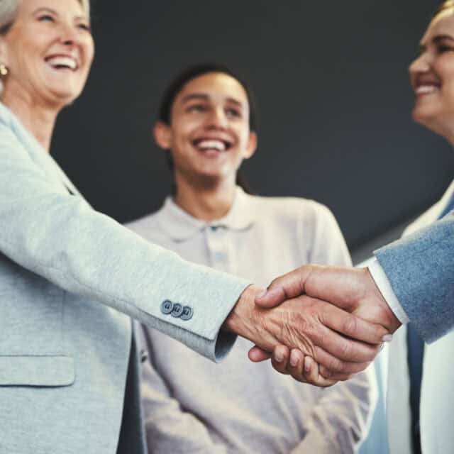 Two professionals shaking hands in a friendly manner to finalize supplier management negotiations, with two smiling colleagues watching, in a well-lit office setting.