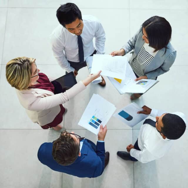 Four business professionals standing, engaged in a discussion with documents and bar charts in hand, demonstrating mobility, viewed from above.