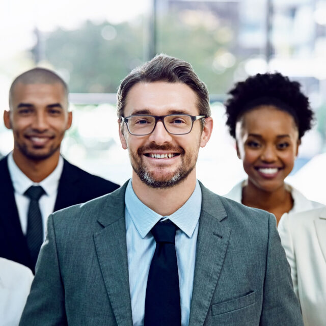 Three business professionals smiling, with a Caucasian man in the foreground and a Black man and a Black woman in the background, standing in an office setting, engaged in project management.