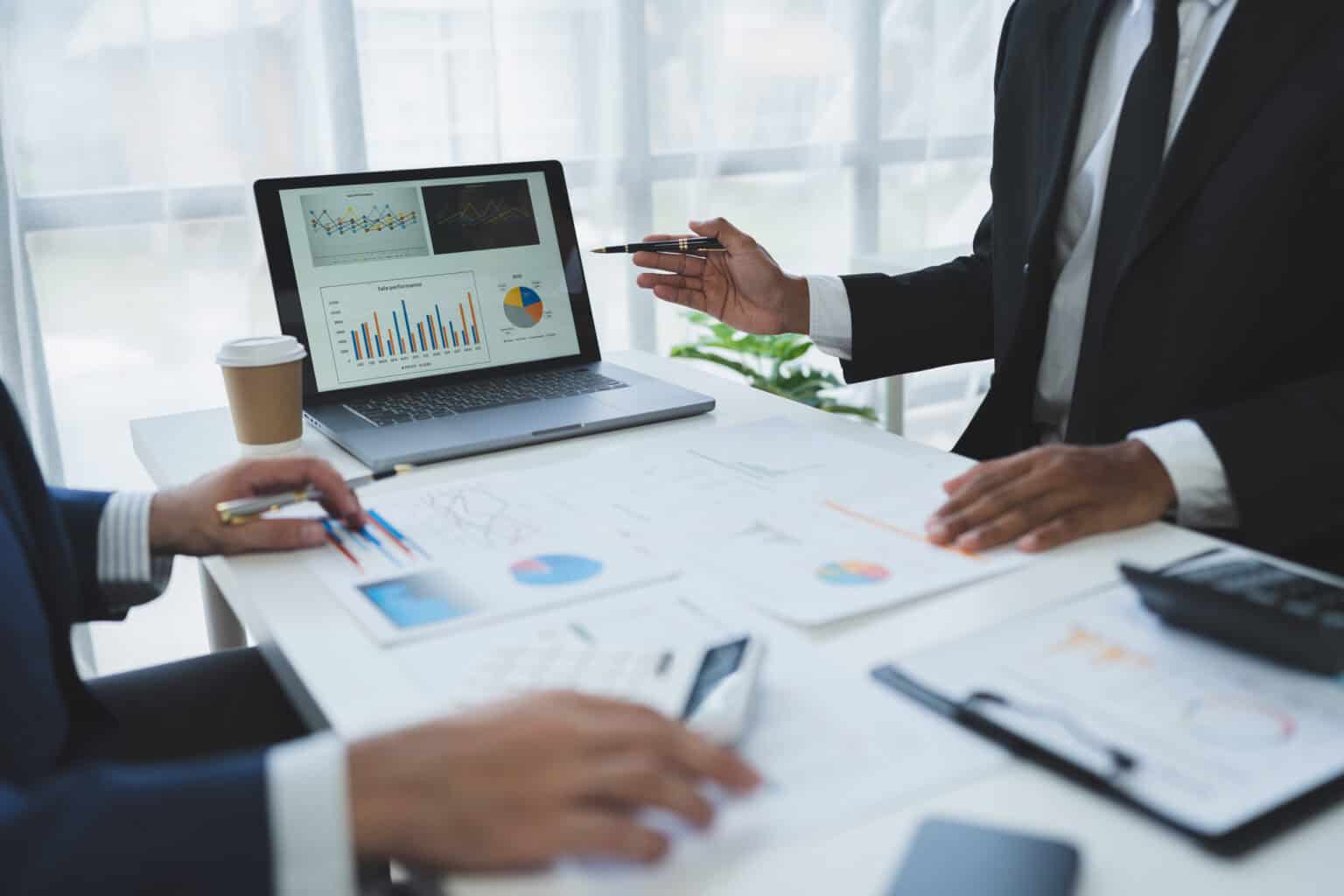 Two people in suits are seated at a well-lit office desk reviewing graphs and charts on paper. One person gestures with a pen, while a laptop displaying data visualizations related to the company's privacy policy is open. A coffee cup and calculator are also on the desk.