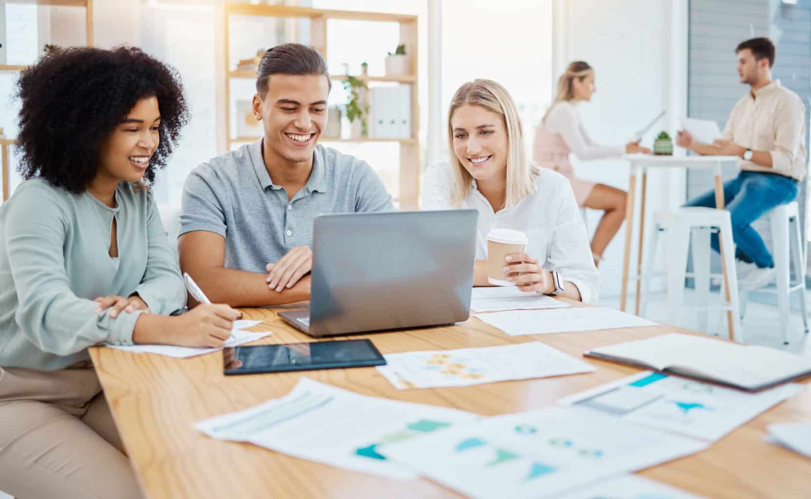 Three colleagues smiling and discussing solutions on a laptop at a modern office table, with others in the background.