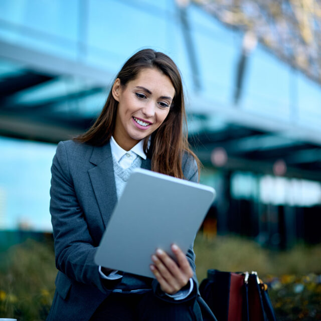 A professional woman in a suit using a tablet outdoors with a smile, focusing on engineering sales, with a modern office building in the background.