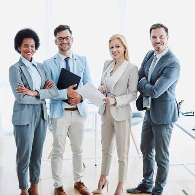 Four business professionals, two men and two women, specializing in supplier management, smiling and standing confidently in a bright office setting.
