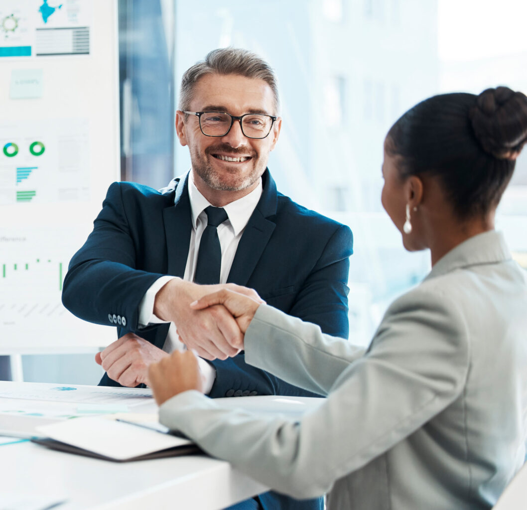 Two professionals shaking hands in an office, smiling, with financial services charts displayed in the background.