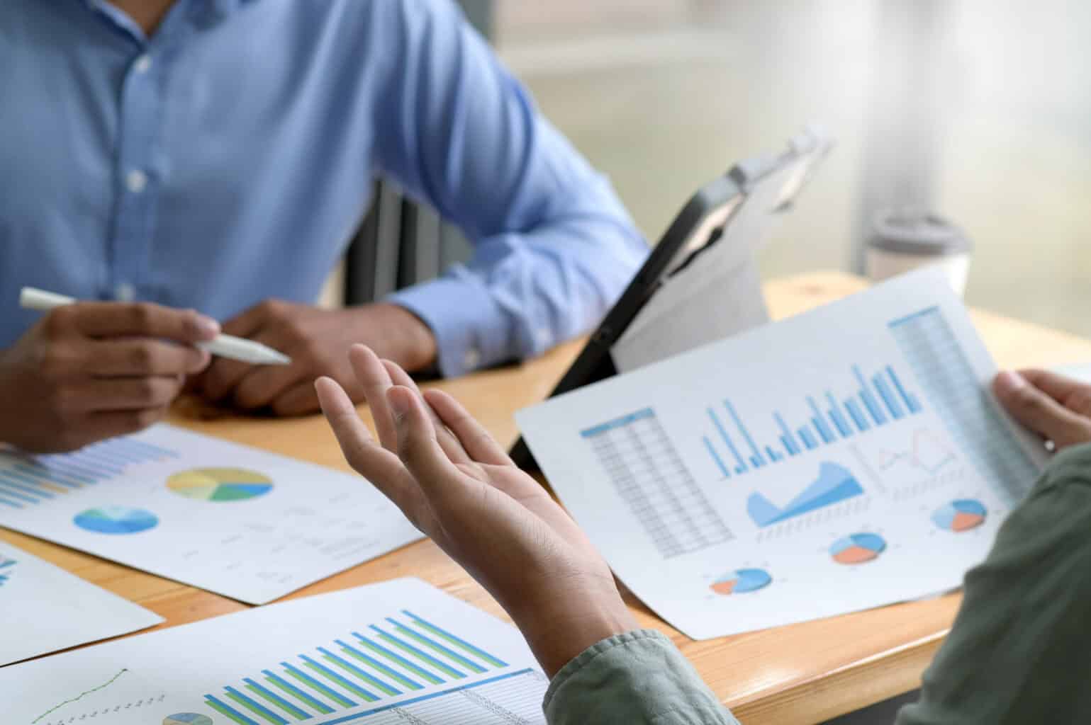 Two individuals are seated at a table reviewing financial charts and graphs. One person points at a chart with a pen, while the other holds a sheet with multiple colorful data visualizations. A laptop displaying the Terms of Service is partially visible on the table.