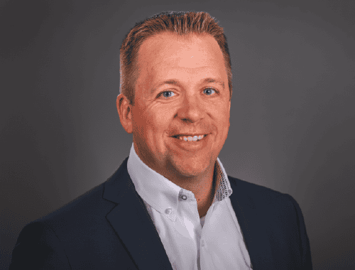 Professional headshot of a smiling man with short blond hair, wearing a blue suit jacket and a white shirt against a gray background.