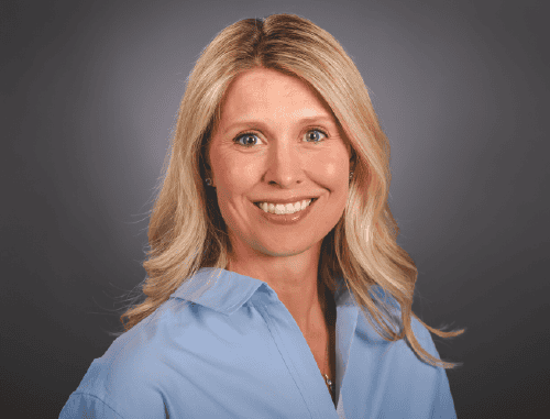 Portrait of a smiling woman with blonde hair, wearing a light blue shirt against a gray background.