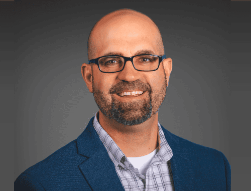 A middle-aged man with a beard and glasses, wearing a blue blazer and a plaid shirt, smiling against a grey background.