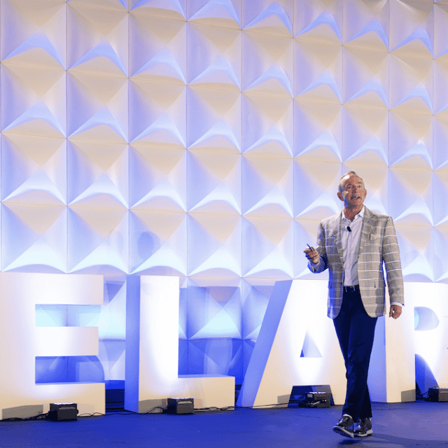 A man in a checked blazer speaks animatedly on stage at a conference with "stellarust" text in the foreground and a geometric patterned backdrop.