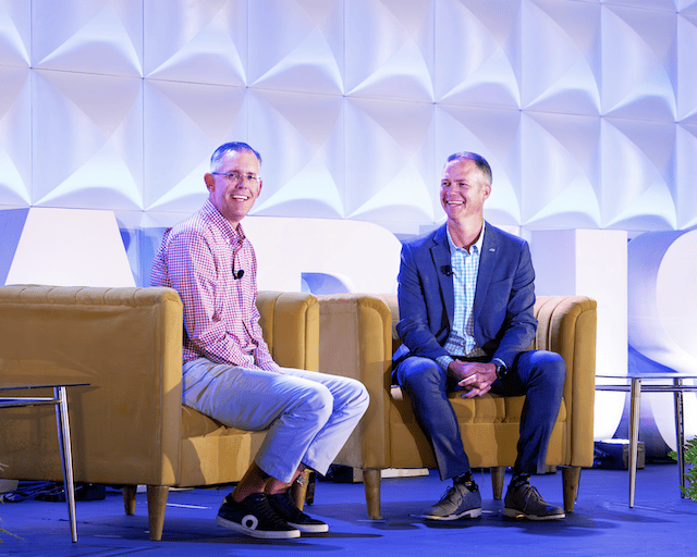 Two men in casual attire smiling and sitting on armchairs on a stage with a geometric patterned backdrop.