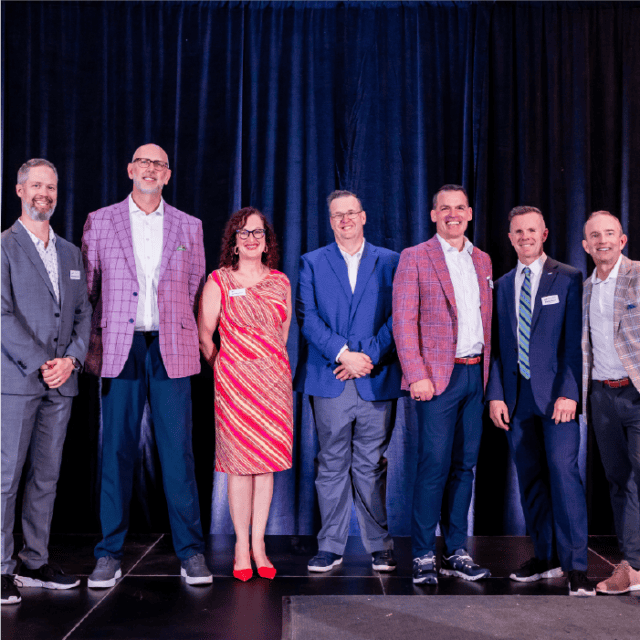 Seven leadership professionals standing on stage, smiling for a group photo at a formal event. They are dressed in smart outfits, including suits and a dress.