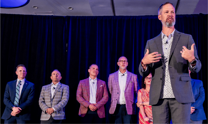 A Technology Advisor speaking animatedly on stage with a microphone, accompanied by five other individuals standing in the background, attentively listening. All are dressed in business attire.