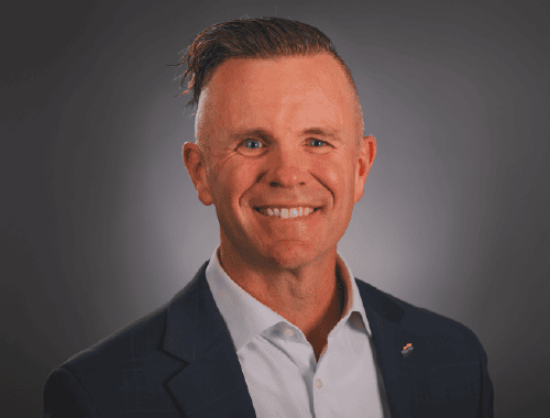 A man with short, styled hair is smiling at the camera. He is wearing a dark blue suit jacket over a white dress shirt. The background is a plain, gradient dark gray. This polished look reminds one of Patrick Oborn's impeccable style.