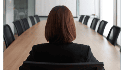 A person with shoulder-length brown hair sits alone at the head of a long conference table in a meeting room, preparing to discuss MSPs buying decisions, while facing a blank screen and a flipchart.