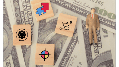 Wooden blocks with business icons and a figurine rest on overlapping $100 bills, evoking themes of strategy, planning, finance, and the dynamics of mergers and acquisitions ahead of Network Shakeup 2025.