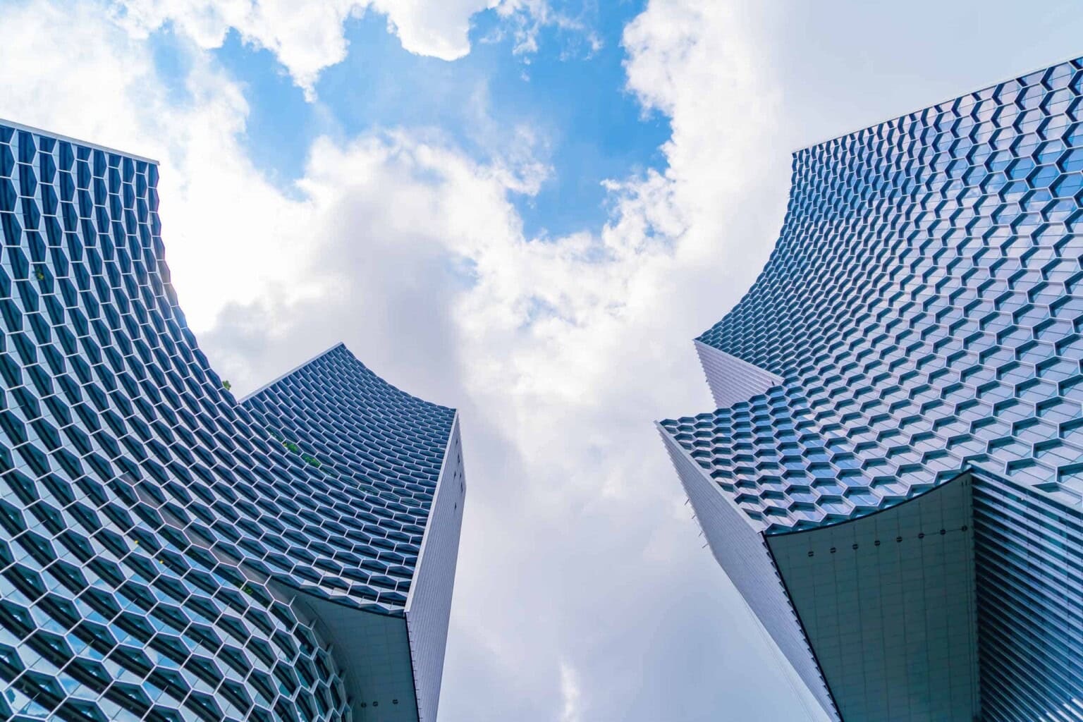 Two modern high-rise buildings with honeycomb-patterned facades curve towards a partly cloudy blue sky, seen from a low-angle perspective.