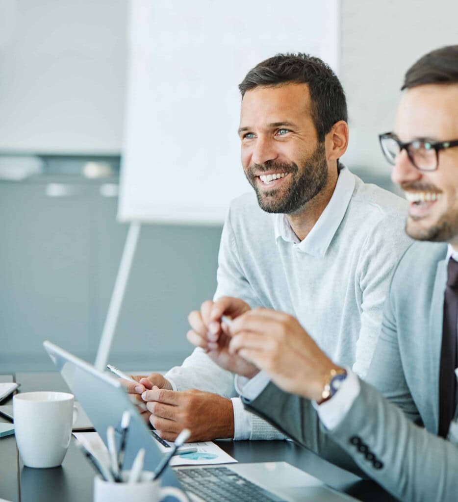 Two men in business attire sit at a desk, smiling and engaged in conversation. One man uses a laptop, and office supplies and a coffee cup are visible on the table. A flip chart is blurred in the background.