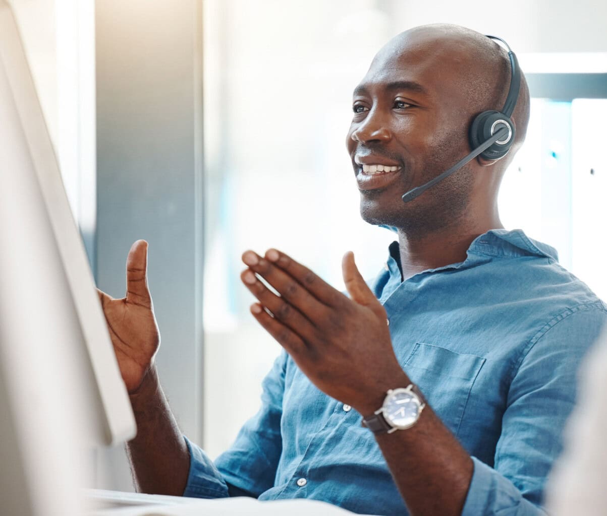 A man wearing a headset and a blue shirt sits at a desk in front of a computer, smiling and gesturing with his hands as if engaged in a video call or customer service conversation.