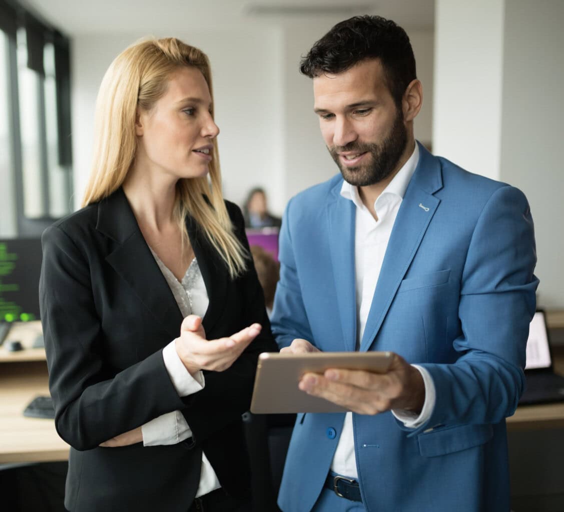 A woman in a black suit and a man in a blue suit are standing in an office, having a discussion while looking at a tablet the man is holding. They appear to be engaged in a professional conversation.