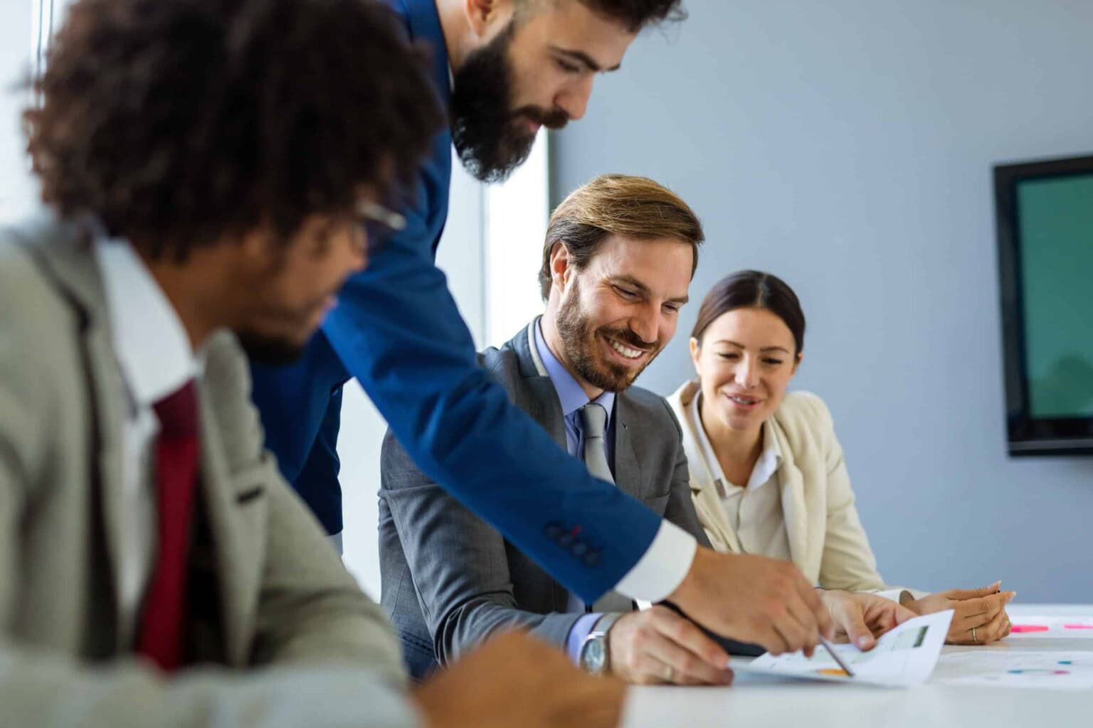 Four business professionals in suits are gathered around a table in a modern office, smiling and discussing documents together. One person is pointing at a paper while the others listen attentively.