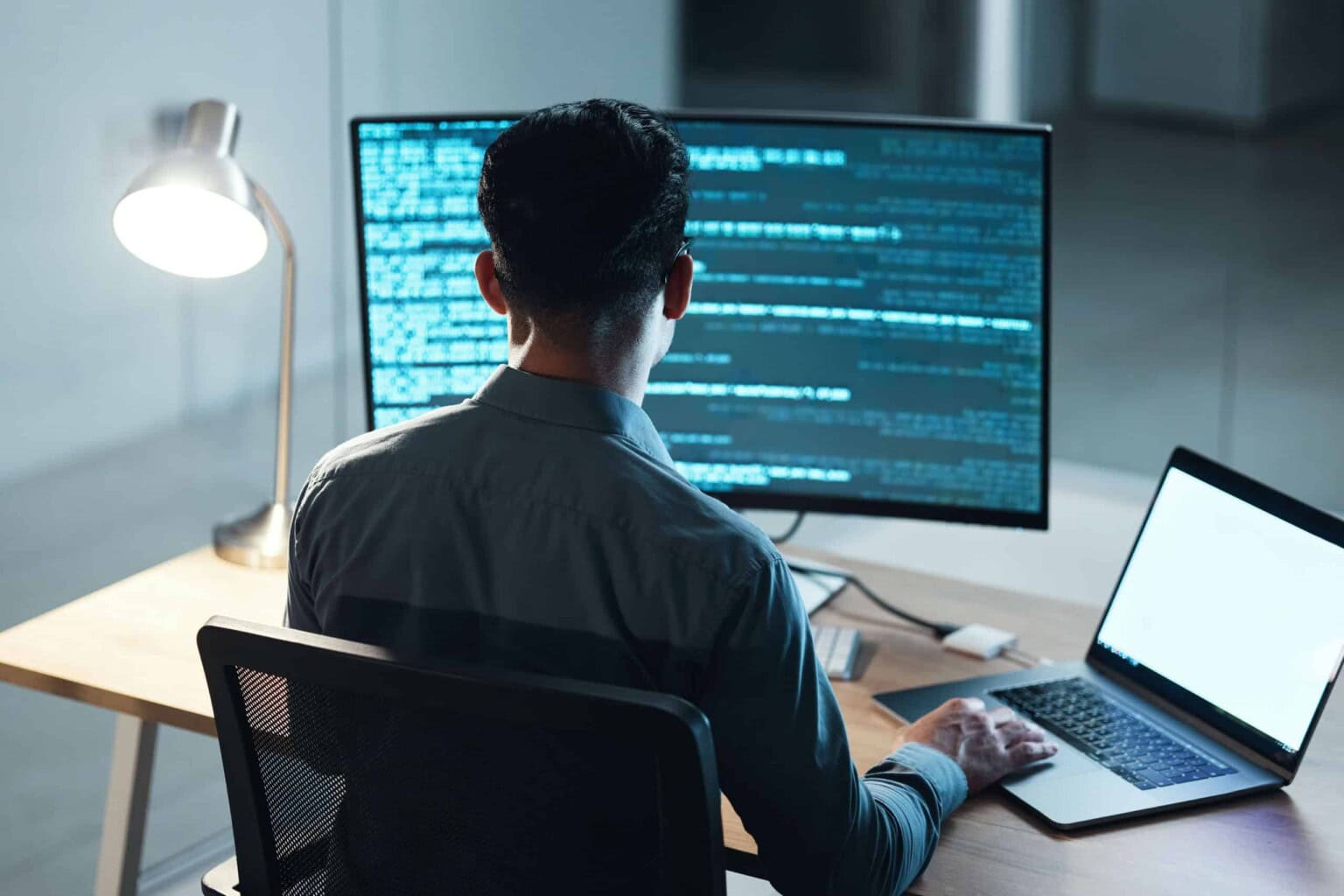 A person sits at a desk working on a computer with code displayed on a large monitor and a laptop. A desk lamp is lit beside them in a modern office setting.