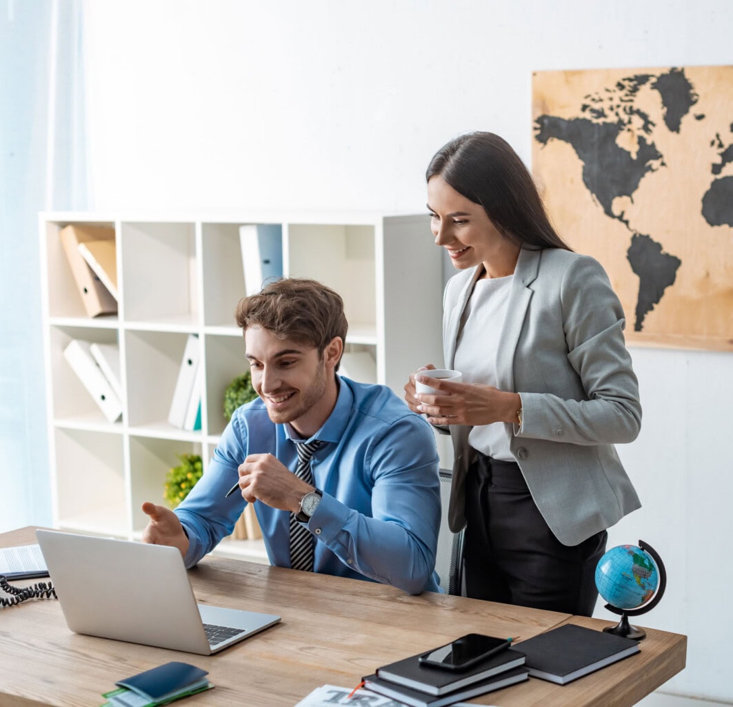 A man in a blue shirt and tie sits at a desk, smiling and looking at a laptop, while a woman in a gray blazer stands beside him holding a cup. Office shelves and a world map are in the background.