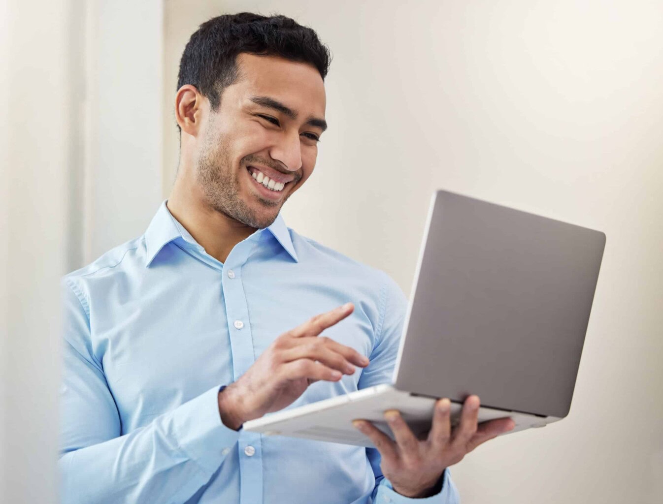 Smiling man in a light blue shirt stands indoors, holding and using a silver laptop computer.