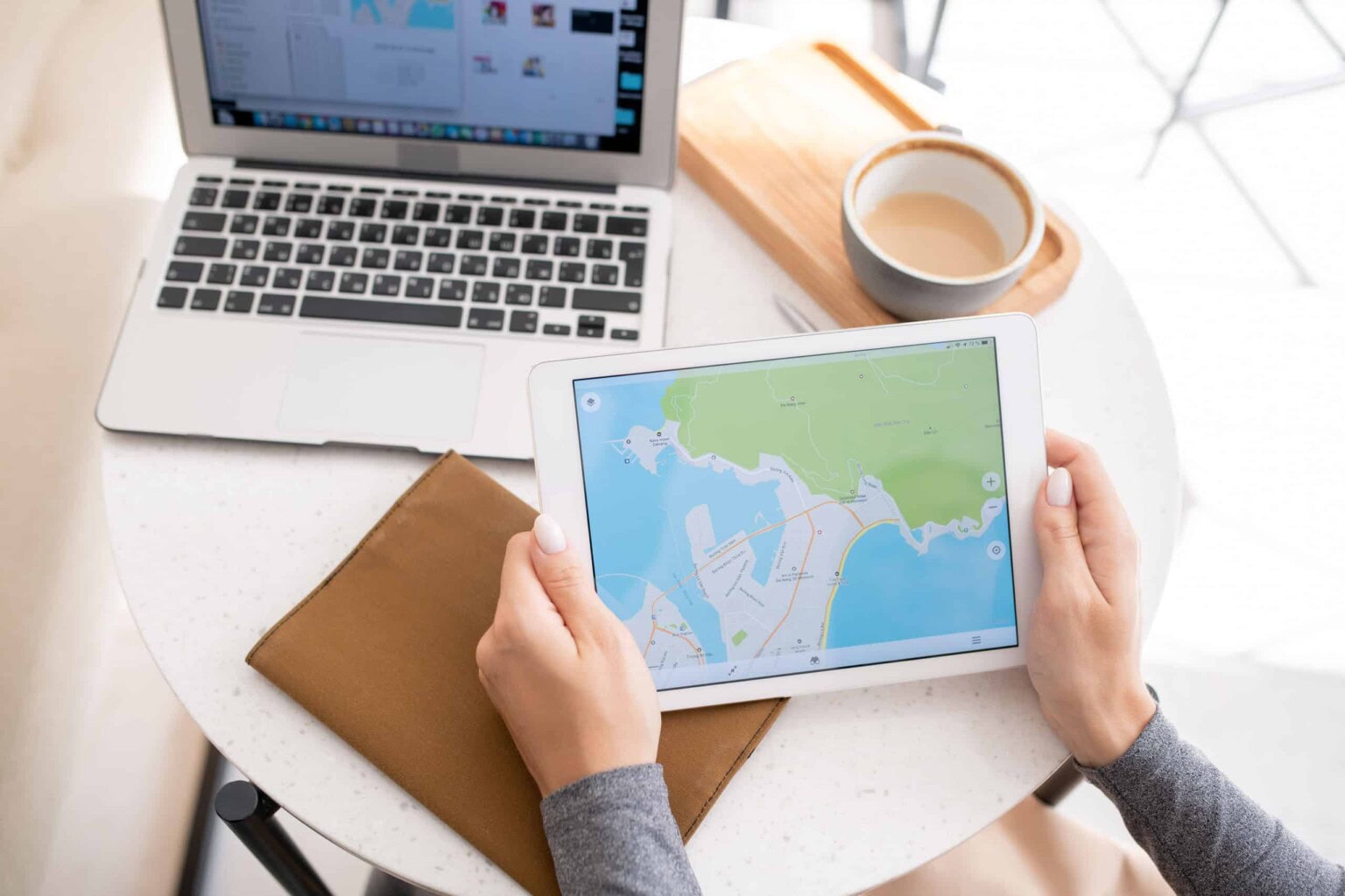 Person holding a tablet displaying a map, sitting at a round table with a laptop, coffee cup, and brown folder.