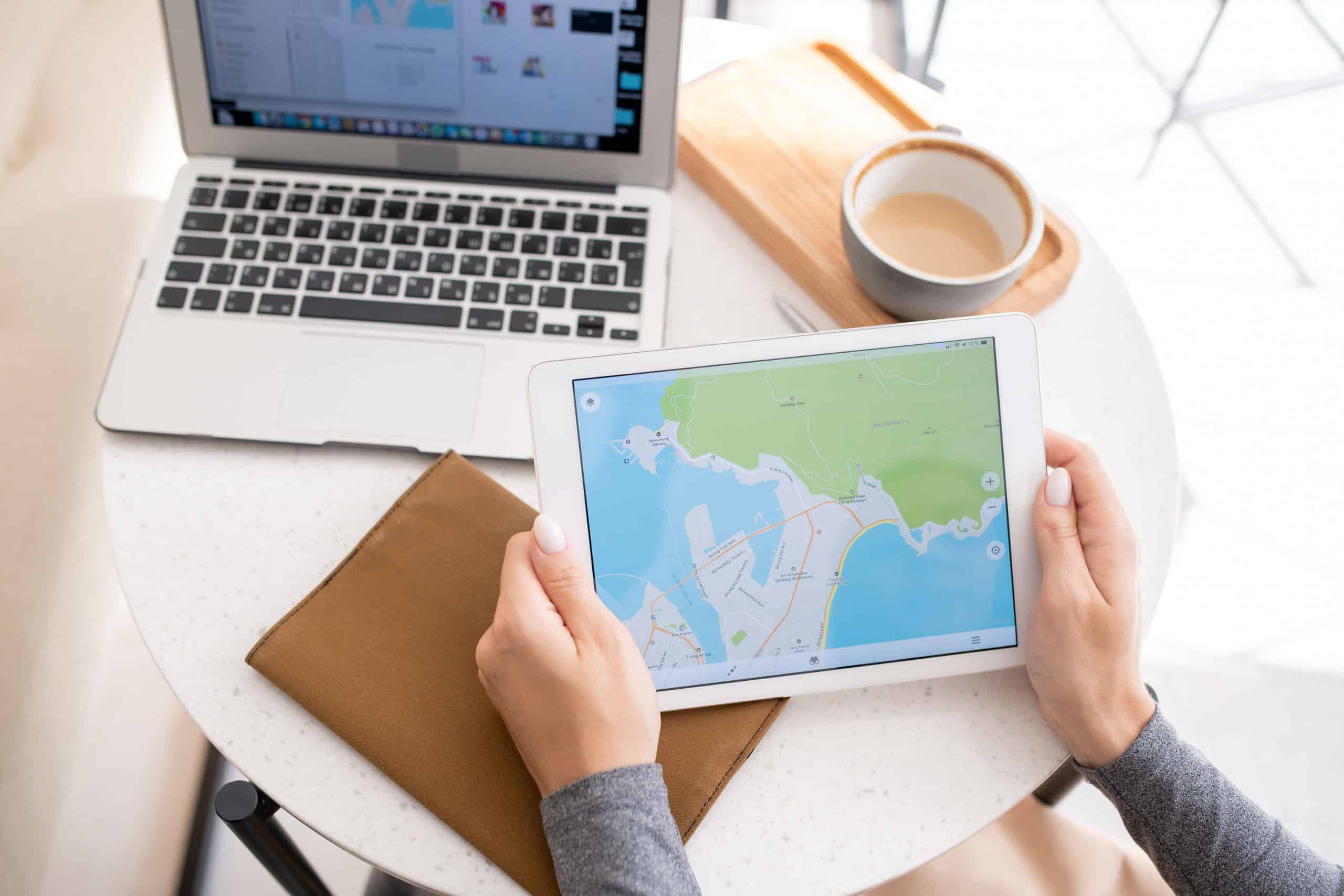 Person holding a tablet displaying a map, sitting at a round table with a laptop, coffee cup, and brown folder.