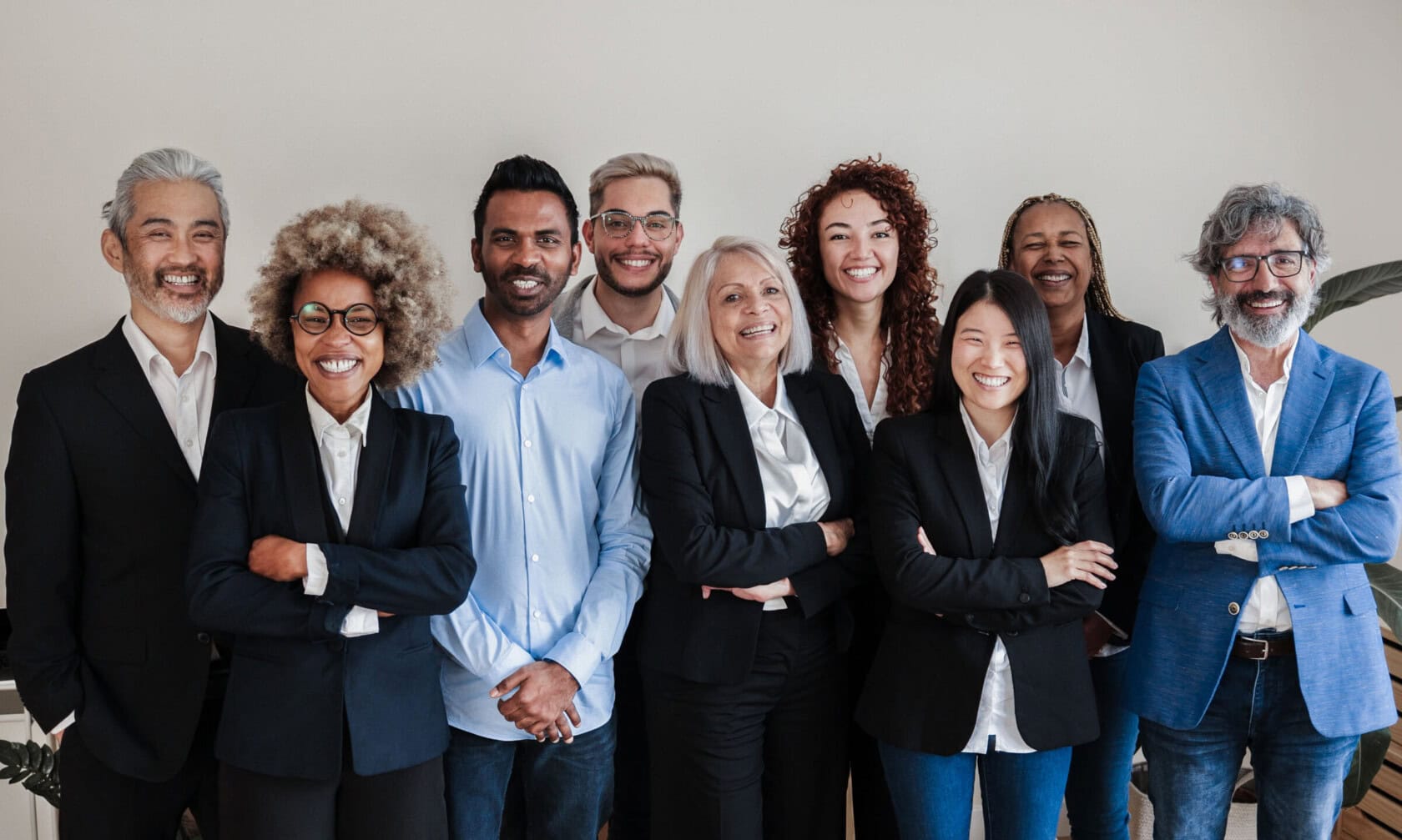 A diverse group of ten professionally dressed people stand in a row, smiling at the camera in a bright indoor setting. Most have their arms crossed, conveying confidence and friendliness.