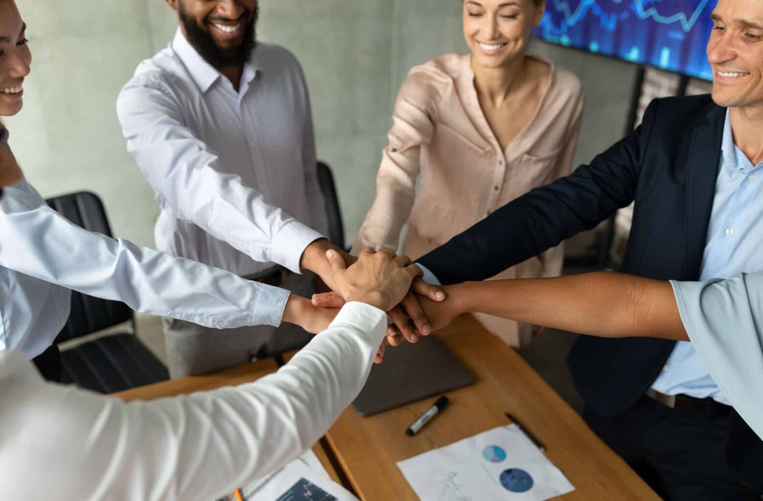 Five business professionals stand around a table, smiling and stacking their hands together in a gesture of teamwork and unity. Documents and a laptop are visible on the table.
