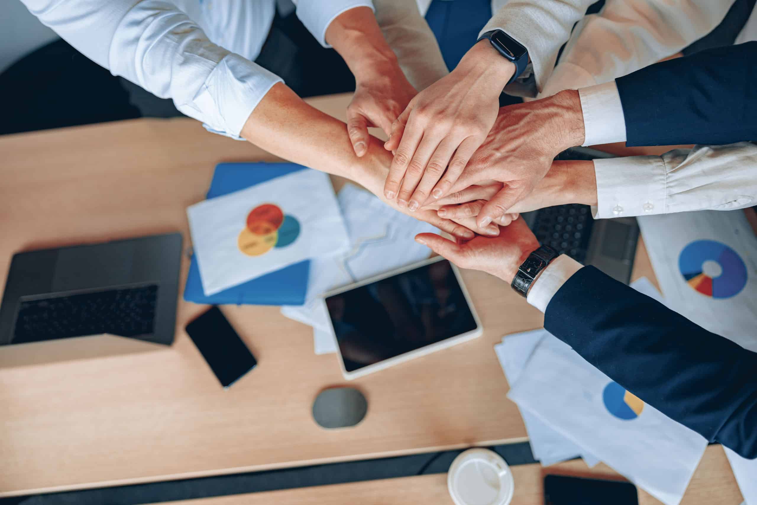 A group of people place their hands together in a teamwork gesture over a desk with laptops, documents, charts, and a tablet. The scene suggests collaboration and a business environment.