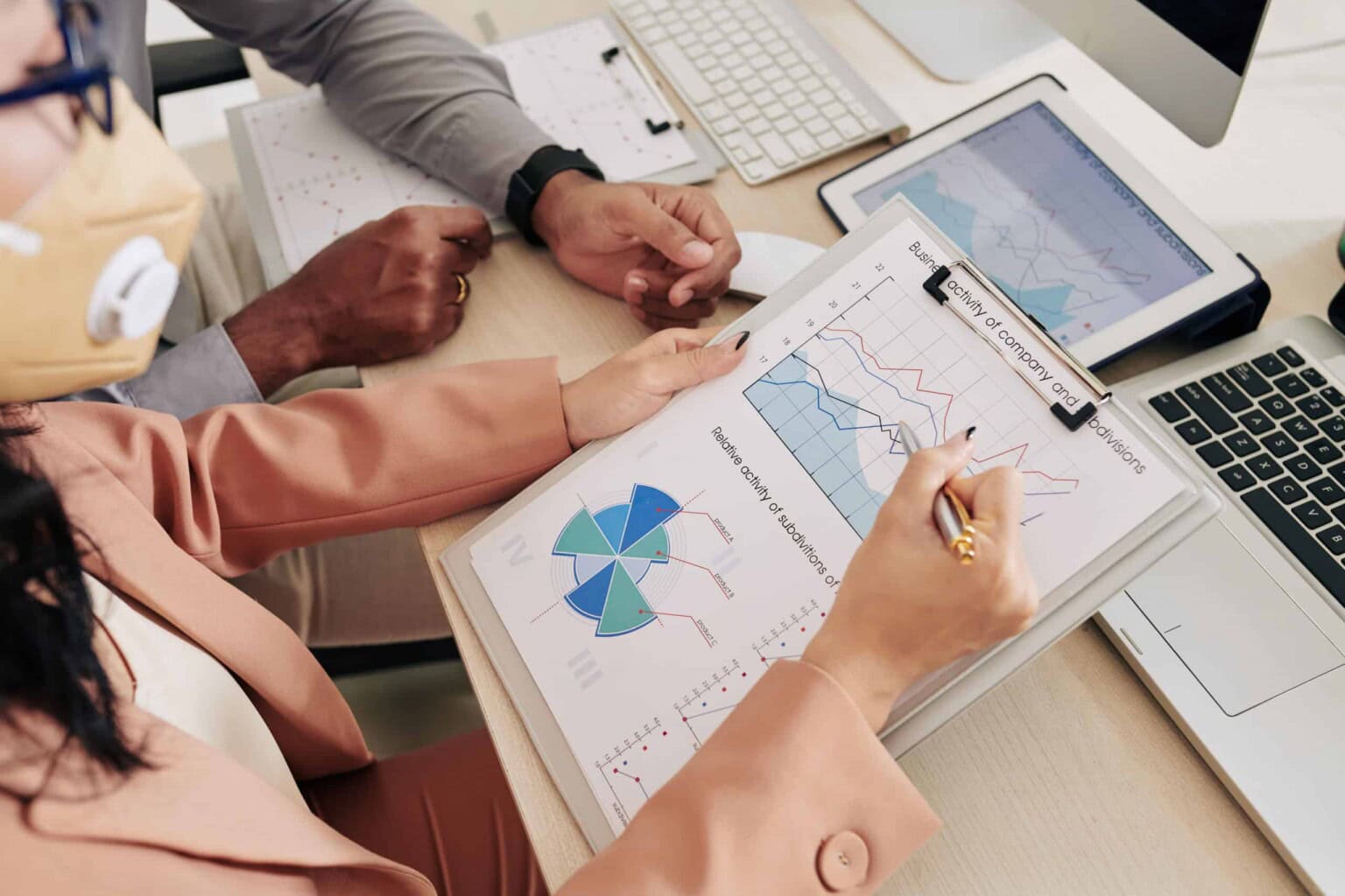 Two people analyzing financial charts and graphs on paper and a tablet at a desk. One person is pointing at a line graph with a pen. Laptops and documents are also on the table.