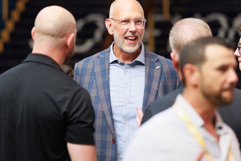 A bald man in a plaid suit jacket and glasses smiles and speaks with several other men at an indoor event. The scene appears casual and conversational.