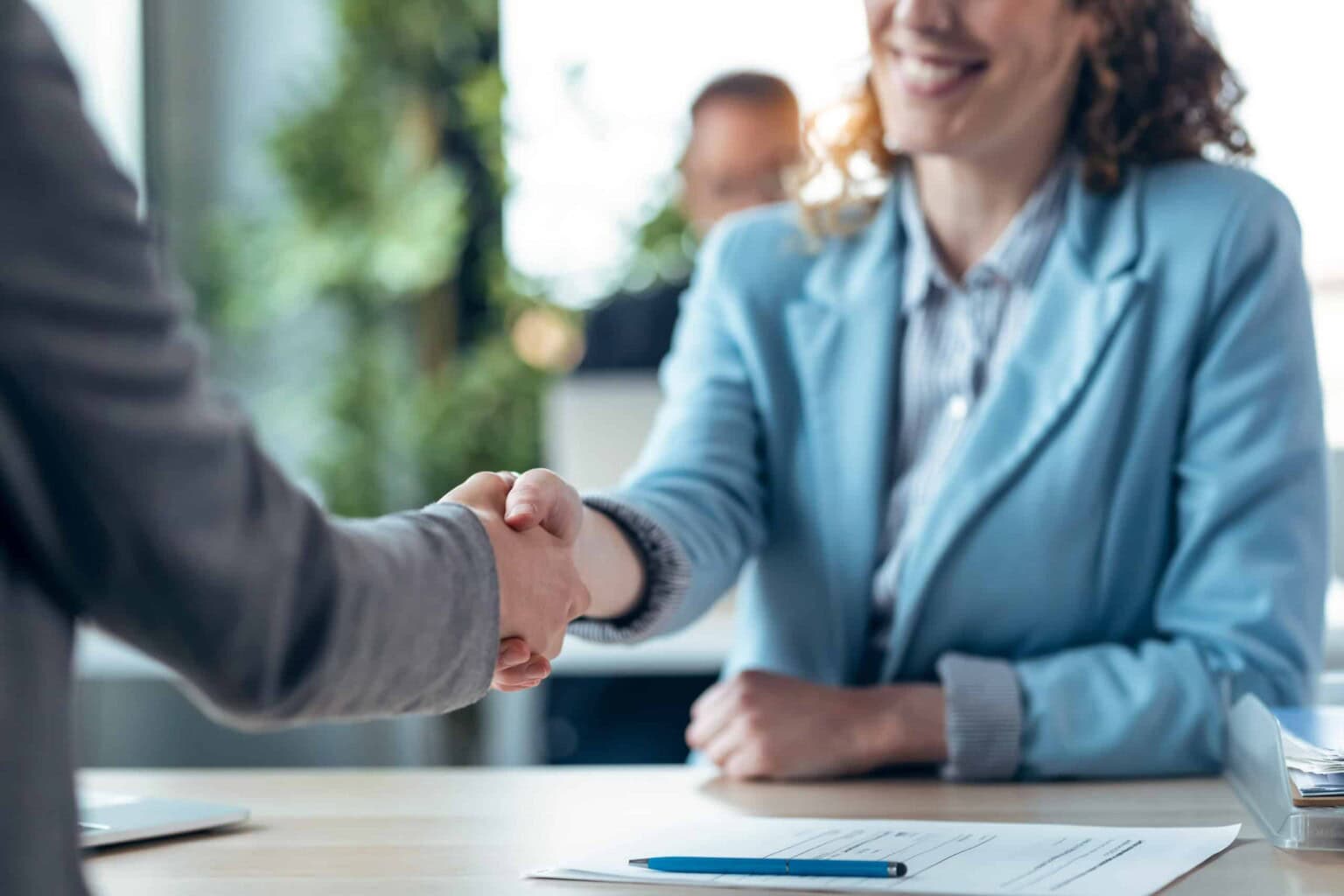 Two people shaking hands across a desk in a professional setting. One person is wearing a light blue blazer and smiling, while documents and a pen are visible on the desk.