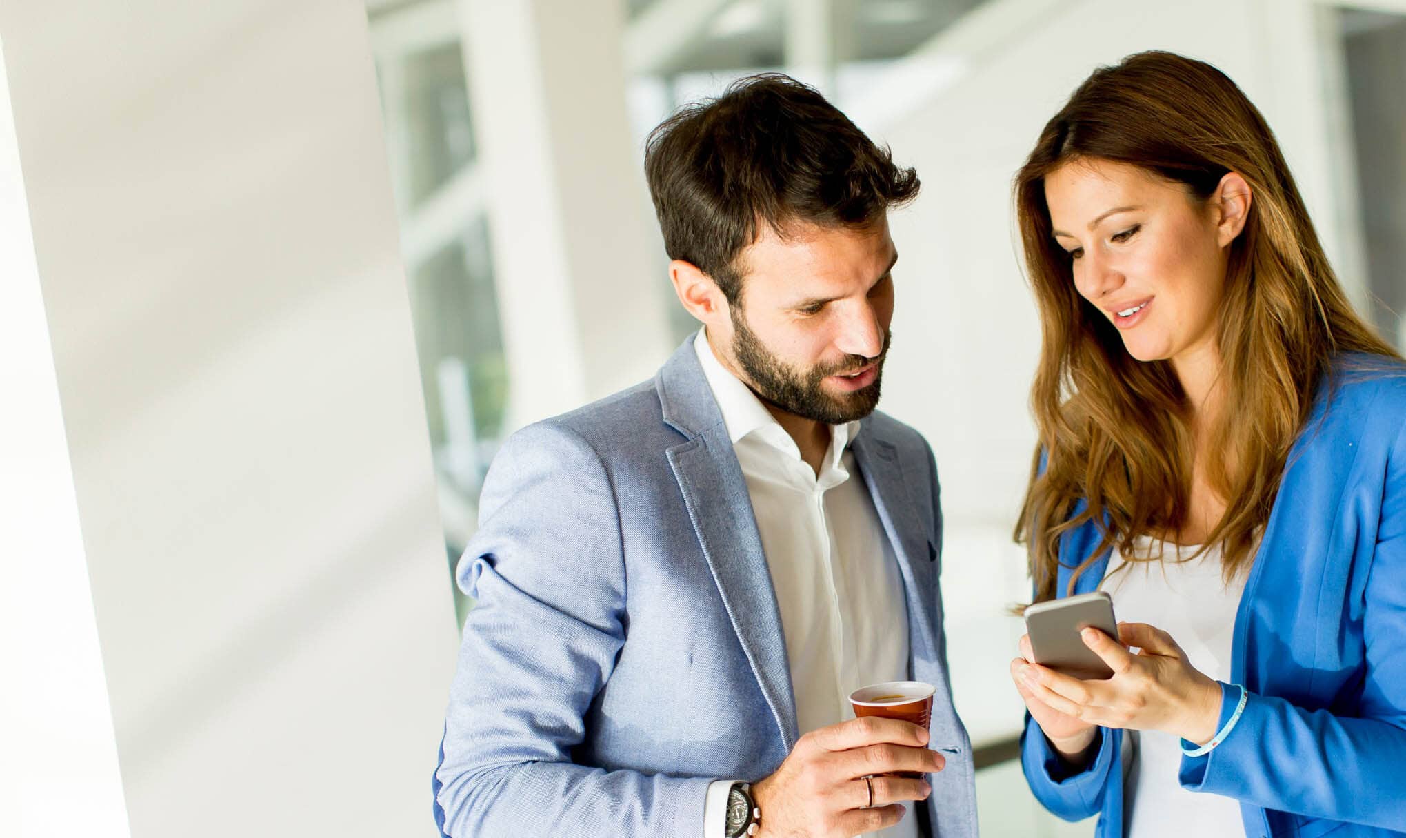 A man and woman in business attire stand indoors. The woman holds a smartphone and smiles while showing it to the man, who holds a small cup and looks at the phone. They appear to be having a friendly conversation.