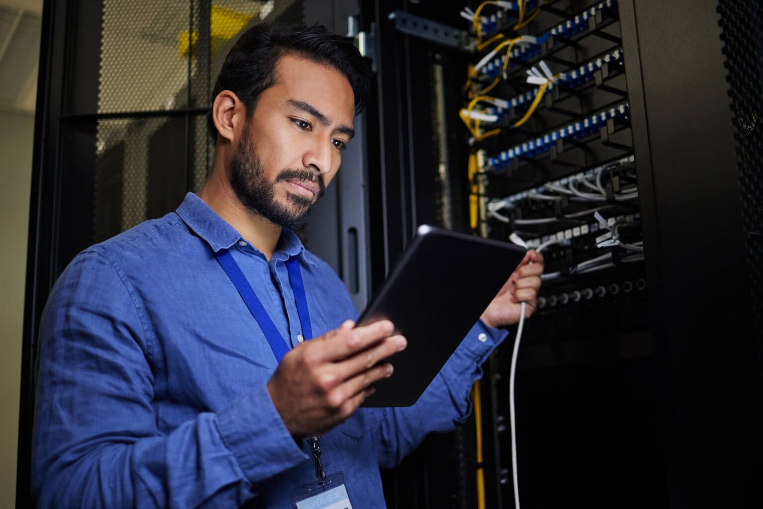 A man in a blue shirt stands in a server room, holding a tablet and a network cable, examining server racks filled with wires and equipment.