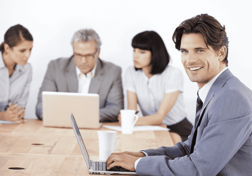 A smiling man in a suit works on a laptop at a conference table, while three colleagues sit in the background having a discussion and using another laptop.