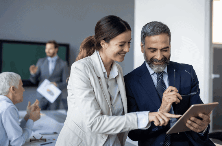A smiling woman and man in business attire look at a tablet together in an office setting, while two other colleagues interact in the background.
