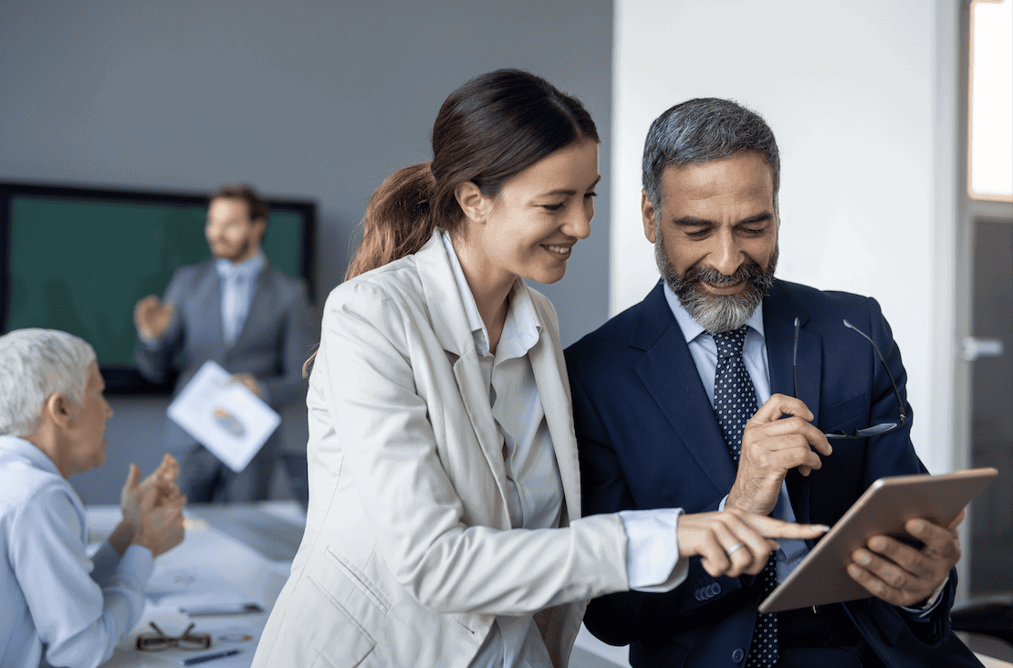 A smiling woman and man in business attire look at a tablet together in an office setting, while two other colleagues interact in the background.