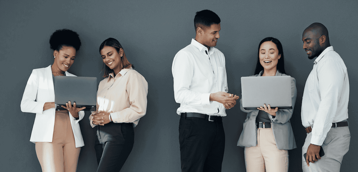 Five professionally dressed people stand against a gray background, interacting in pairs and using laptops while smiling and engaging in conversation.