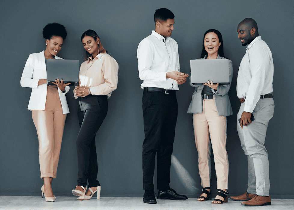 Five professionally dressed people stand against a gray wall, smiling and interacting while holding laptops, suggesting a collaborative and friendly work environment.