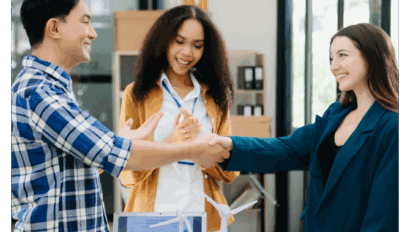 Three colleagues in an office environment, two shaking hands and smiling while a third person stands between them, also smiling. A computer with charts is visible on the desk in the foreground.