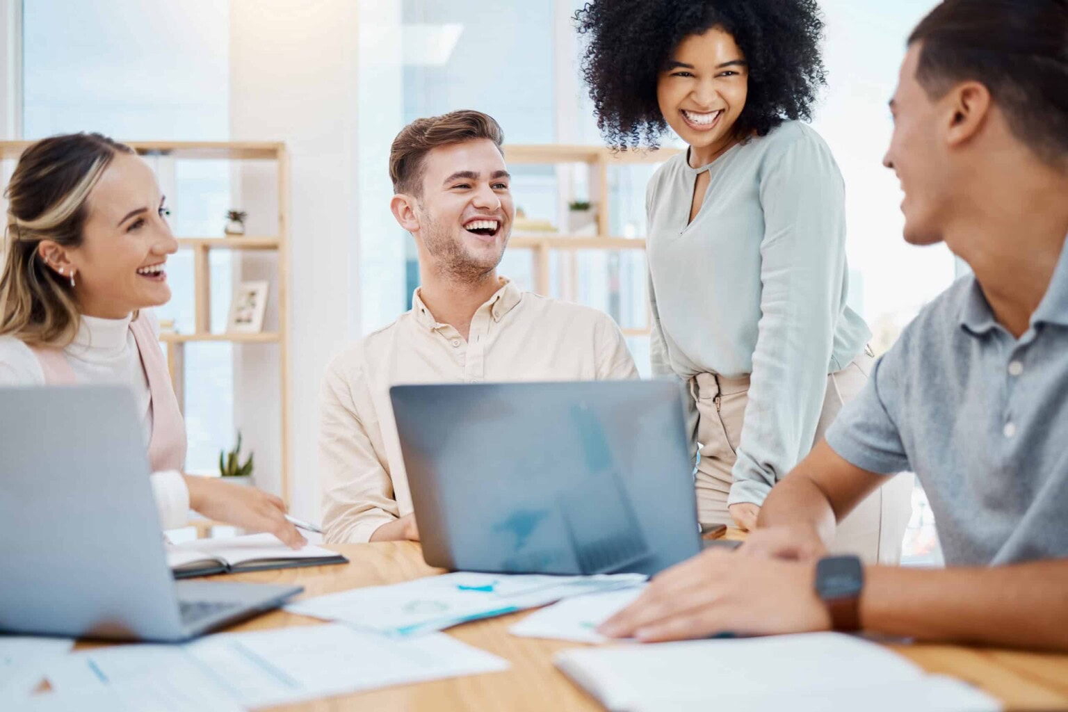 Four young professionals sit around a table with laptops and papers, smiling and laughing together in a bright office setting, suggesting a collaborative and positive work environment.