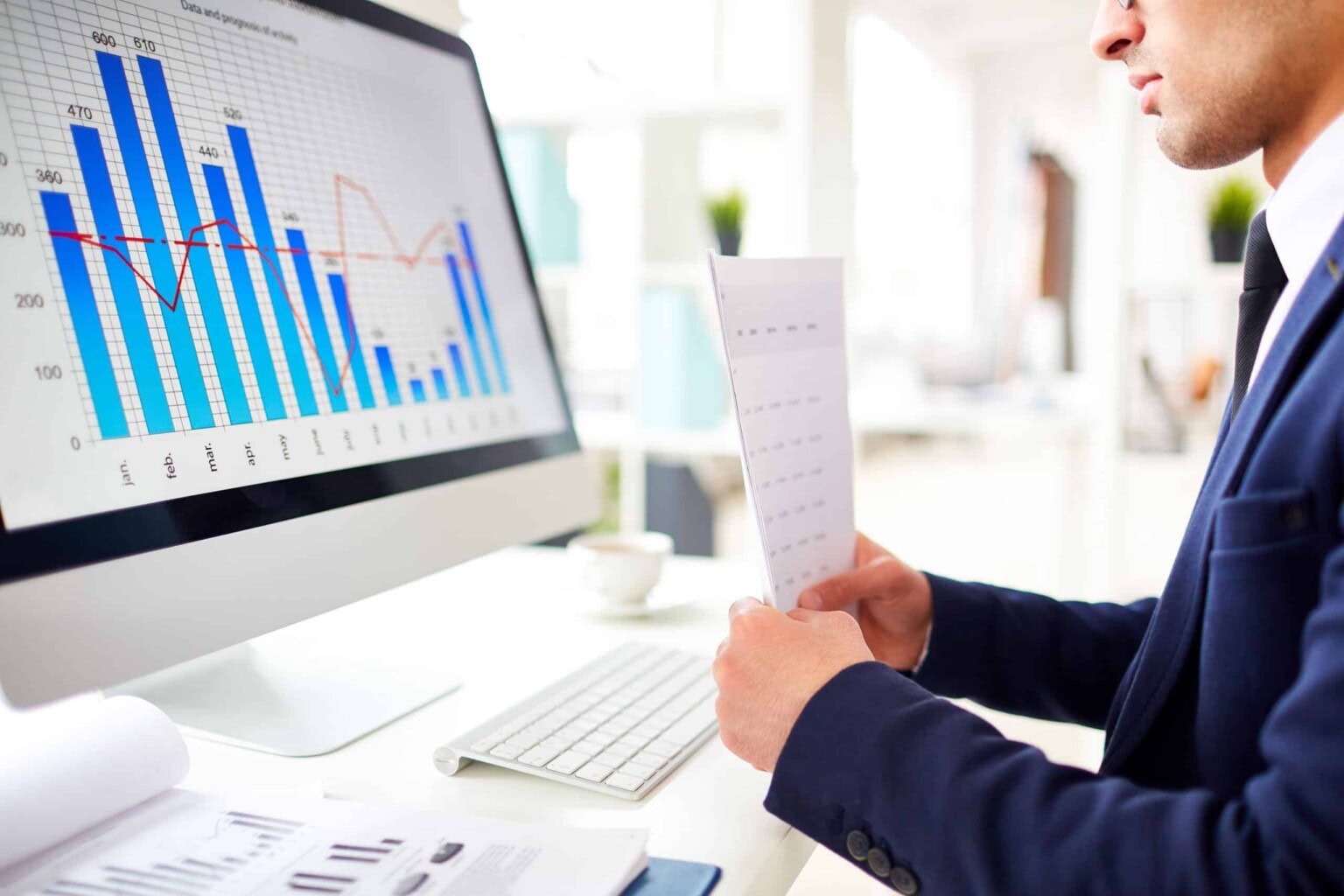 A person in a suit sits at a desk, holding papers and analyzing a computer screen displaying bar and line graphs with financial data. A keyboard, mouse, and documents are on the desk.