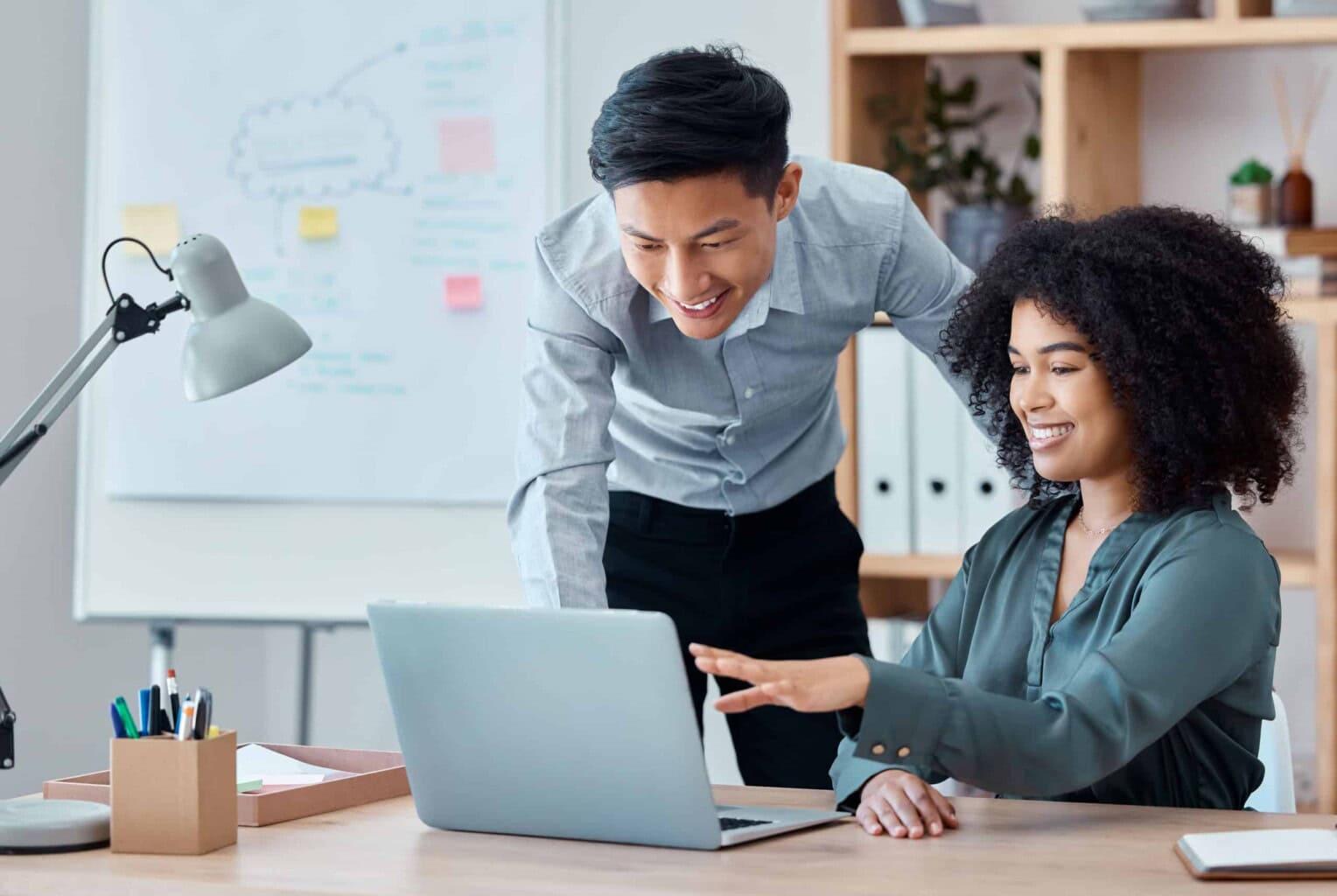 Two colleagues, a man and a woman, smile while looking at a laptop screen in a bright office. The woman is seated and pointing at the screen; the man stands beside her, leaning in. Shelves and a whiteboard are visible in the background.