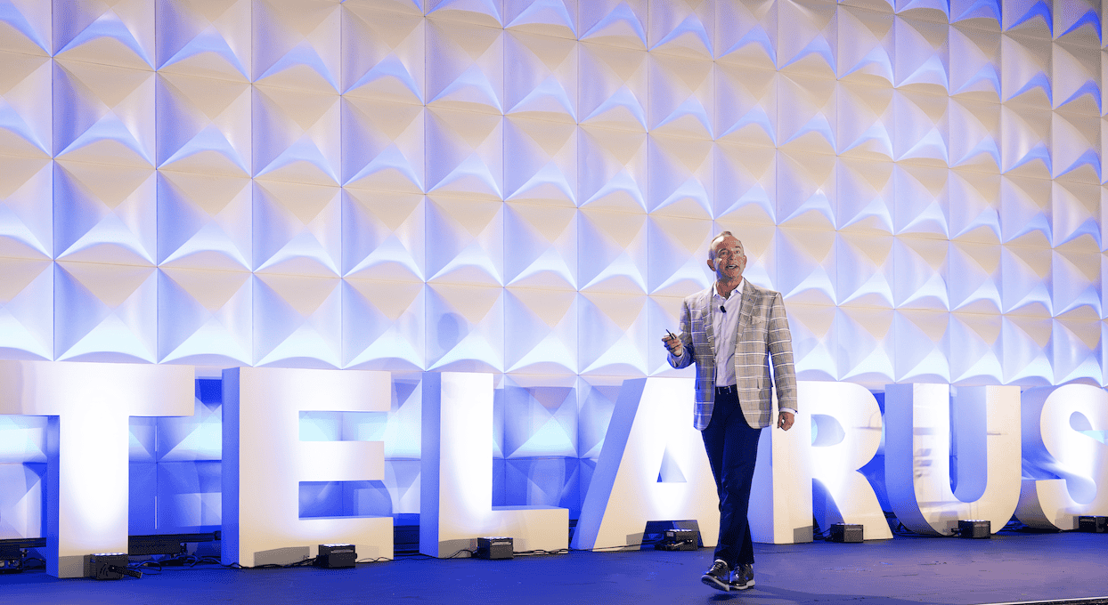 A man in a plaid blazer stands on a stage, holding a microphone, in front of large illuminated letters spelling "TELARUS" against a geometric, blue-lit backdrop.